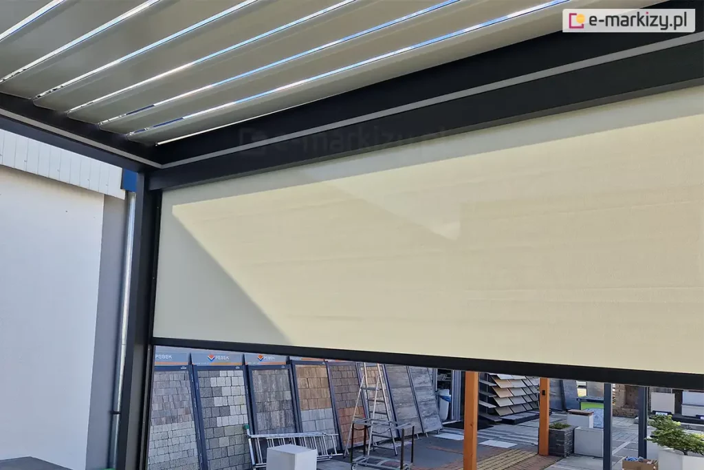 Widok z wnętrza nowoczesnej pergoli tarasowej na system bocznych osłon przeciwsłonecznych typu screen. Na pierwszym planie widoczny jest refleksol Ziiip 120 Selt w kolorze beżowym, który rozwinięty jest na część wysokości, stanowiąc skuteczną barierę przed słońcem i wiatrem. Tkanina charakteryzuje się gładkim splotem i jest stabilnie zamontowana w antracytowej ramie konstrukcyjnej pergoli. Powyżej rolety widoczne jest zadaszenie pergoli wykonane z ruchomych, aluminiowych lameli (system bioklimatyczny) w kolorze białym, ustawionych pod kątem, co pozwala na regulację dopływu światła. W tle, za konstrukcją pergoli, znajduje się ekspozycja zewnętrzna z różnymi rodzajami kostki brukowej, płyt tarasowych oraz elementami małej architektury ogrodowej. Całość prezentuje funkcjonalne wykorzystanie rolet screen jako integralnego elementu nowoczesnych systemów zadaszeń tarasowych.