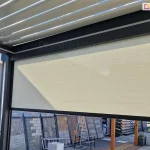 Widok z wnętrza nowoczesnej pergoli tarasowej na system bocznych osłon przeciwsłonecznych typu screen. Na pierwszym planie widoczny jest refleksol Ziiip 120 Selt w kolorze beżowym, który rozwinięty jest na część wysokości, stanowiąc skuteczną barierę przed słońcem i wiatrem. Tkanina charakteryzuje się gładkim splotem i jest stabilnie zamontowana w antracytowej ramie konstrukcyjnej pergoli. Powyżej rolety widoczne jest zadaszenie pergoli wykonane z ruchomych, aluminiowych lameli (system bioklimatyczny) w kolorze białym, ustawionych pod kątem, co pozwala na regulację dopływu światła. W tle, za konstrukcją pergoli, znajduje się ekspozycja zewnętrzna z różnymi rodzajami kostki brukowej, płyt tarasowych oraz elementami małej architektury ogrodowej. Całość prezentuje funkcjonalne wykorzystanie rolet screen jako integralnego elementu nowoczesnych systemów zadaszeń tarasowych.