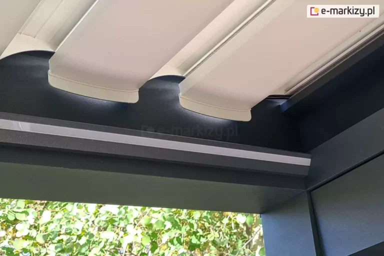 Zbliżenie na narożny detal konstrukcyjny nowoczesnej pergoli bioklimatycznej w miejscu połączenia ruchomego dachu z ramą nośną. Szerokie, aluminiowe lamele w kolorze białym są ustawione w pozycji lekko otwartej, ukazując precyzyjnie wyprofilowane zakończenia z białego tworzywa, które płynnie nachodzą na czarną szynę prowadzącą. Główna belka konstrukcyjna oraz masywny słup wykonane są z aluminium malowanego proszkowo na kolor matowego antracytu. Bezpośrednio pod górną krawędzią ramy zamontowana jest liniowa listwa oświetlenia LED, osłonięta mlecznym kloszem, która biegnie wzdłuż całej długości profilu. W tle, pod zadaszeniem, widoczne są gęste korony drzew o zielonych liściach, a ujęcie z dołu podkreśla czystość formy, techniczne spasowanie elementów oraz kontrast między jasnymi lamelami a ciemnym stelażem.