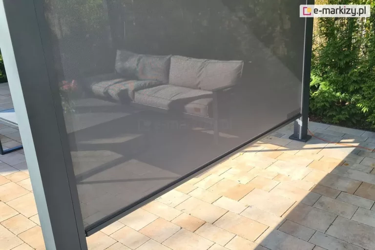 Widok zewnętrzny na niemal całkowicie rozwinięty refleksol Zip z tkaniną ekranową typu screen, zamontowaną w konstrukcji nowoczesnej pergoli tarasowej. Tkanina techniczna w kolorze ciemnoszarym charakteryzuje się drobnym splotem, który mimo dużego stopnia zaciemnienia i ochrony przed słońcem, pozostaje półprzezroczysty, pozwalając na wgląd do wnętrza strefy wypoczynkowej. Za rozwiniętą przesłoną widoczna jest sofa ogrodowa z szarymi poduszkami oraz fragment stolika. Dolna krawędź rolety wykończona jest czarną listwą obciążającą, która porusza się w bocznych prowadnicach zintegrowanych z masywnymi, antracytowymi słupami pergoli. Konstrukcja jest posadowiona na tarasie wyłożonym prostokątną kostką brukową w ciepłych odcieniach beżu i piaskowca, na której widoczne są geometryczne cienie rzucane przez konstrukcję. W tle, po prawej stronie, znajduje się gęsta ściana zielonych krzewów ogrodowych, a naturalne, dzienne światło podkreśla matową teksturę aluminiowych profili.