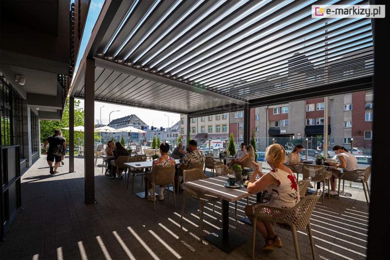 Delikatnie otwarte lamele pergoli wolnostojącej Sb400 wpuszczają odpowiednią ilość promieni słonecznych, by zapewnić klientom restauracji dostęp do naturalnego światła bez narażania na palące promienie słońca. Z zacienionej przestrzeni tarasu na świeżym powietrzu korzysta wielu klientów restauracji. W pobliżu widoczny blok mieszkalny oraz skrzyżowanie z sygnalizacją świetlną.