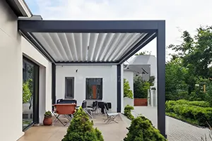 Nowoczesna pergola tarasowa o konstrukcji aluminiowej w kolorze ciemnego antracytu, zamontowana bezpośrednio do jasnej elewacji współczesnego budynku mieszkalnego. Zadaszenie pergoli składa się z poziomych, ruchomych lameli w kolorze białym, które ułożone są w seryjne, równoległe pasy tworzące estetyczną, harmonijkową strukturę osłaniającą taras przed słońcem. Konstrukcja wsparta jest na pionowych, masywnych słupach nośnych o kwadratowym przekroju, które stabilizują narożnik zadaszenia od strony ogrodu. Pod pergolą, na wybrukowanej jasnymi płytami nawierzchni, znajduje się strefa wypoczynkowa z zestawem mebli ogrodowych, w tym z krzesłami turystycznymi, stołem oraz brązowym fotelem. W tle widoczne są białe ściany budynku z ciemnymi ramami okiennymi i dużymi przeszkleniami tarasowymi. Przestrzeń wokół tarasu została zagospodarowana niską roślinnością ogrodową, formowanymi krzewami iglastymi na pierwszym planie, zielonym trawnikiem oraz ułożoną z kostki ścieżką prowadzącą w głąb ogrodu z gęstymi drzewami pod jasnym niebem.