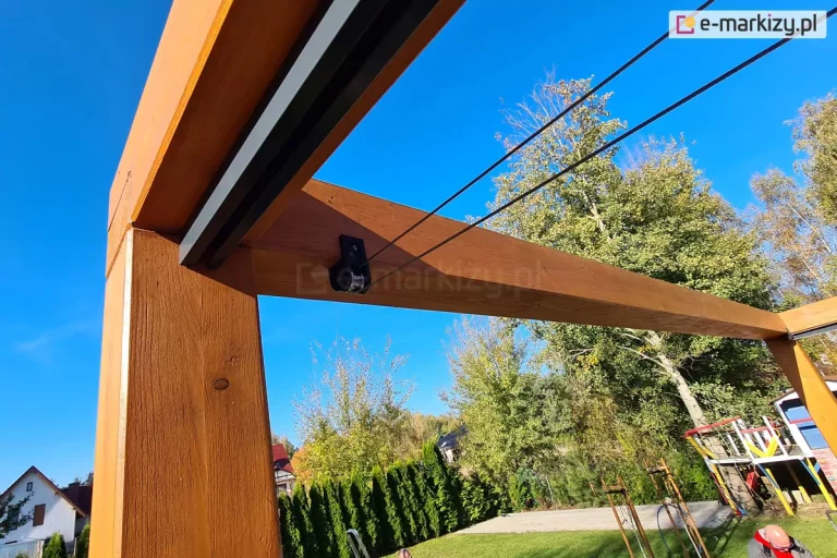 Pergola Koro z dachem Supro mocowanym pod belką z widocznym elementem napędu ręcznego sznurkowego. W tle ogród z trawnikiem i miejscem wyłożonym kostką oraz placem zabaw dla dzieci. W tle drzewa, żywopłot i błękitne niebo.