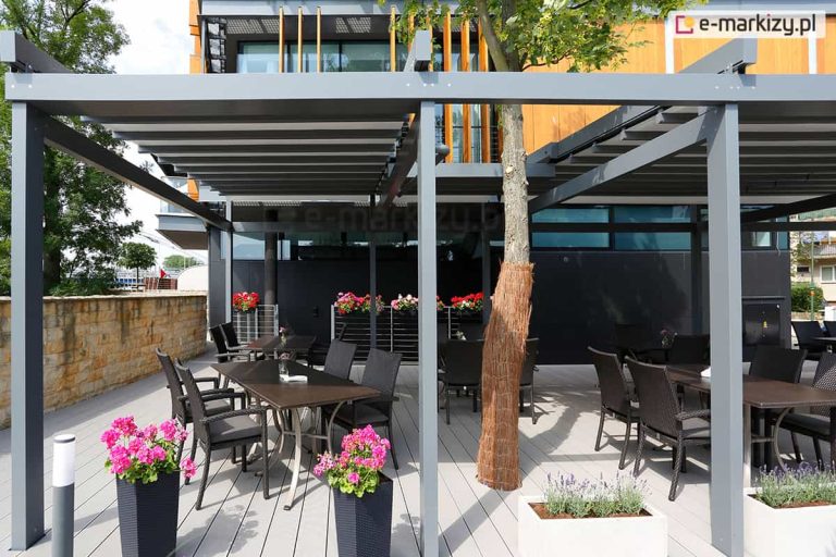 Nowoczesny ogródek restauracyjny osłonięty wielomodułową pergolą tarasową Solid o konstrukcji aluminiowej w kolorze antracytowym. Zadaszenie składa się z szarego, segmentowego materiału. Pod pergolą, na jasnym tarasie z desek kompozytowych, ustawiono ciemne stoły i technorattanowe fotele. Konstrukcja została wkomponowana w otoczenie, uwzględniając rosnące drzewo liściaste, którego pień owinięto brązową matą ochronną. Przestrzeń udekorowana jest licznymi kwiatami: różowymi pelargoniami w wysokich antracytowych donicach oraz lawendą w białych, prostokątnych skrzyniach. W tle widoczny jest nowoczesny budynek z elewacją łączącą czarne panele z pionowymi drewnianymi lamelami oraz kamienny murek po lewej stronie. Całość oświetlona jest naturalnym, dziennym światłem.