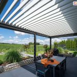 Antracytowa konstrukcja pergoli z jasnymi lamelami przeciwsłonecznymi nad stołem z pomarańczową serwetą i bukietem kwiatów. Doskonale zagospodarowany taras otacza zieleń trawnika oraz liczne rośliny ozdobne