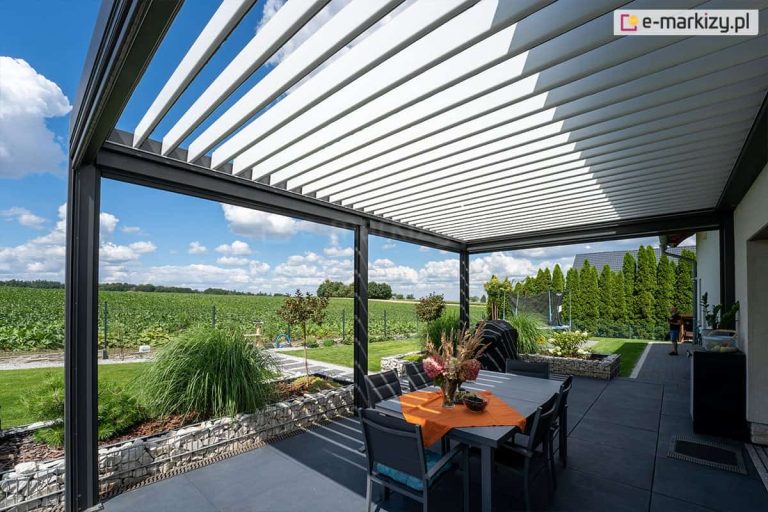 Antracytowa konstrukcja pergoli z jasnymi lamelami przeciwsłonecznymi nad stołem z pomarańczową serwetą i bukietem kwiatów. Doskonale zagospodarowany taras otacza zieleń trawnika oraz liczne rośliny ozdobne.