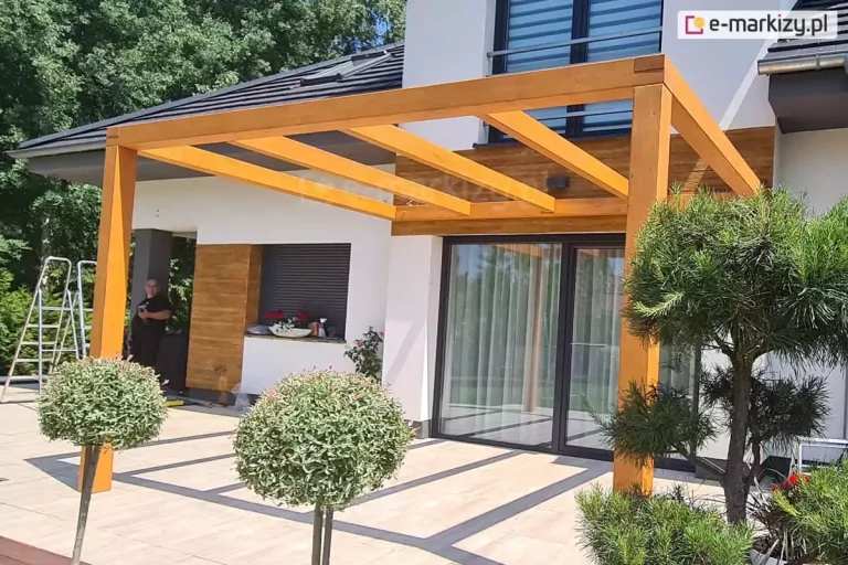 Drewniana przyścienna pergola zamocowana nad drzwiami tarasowymi do elewacji budynku o jasnej fasadzie. Przed tarasem widoczne drzewko iglaste i dwa drzewka liściaste o kulistej koronie. W tle na tarasie stoi mężczyzna, obok którego ustawiono dwie drabiny.