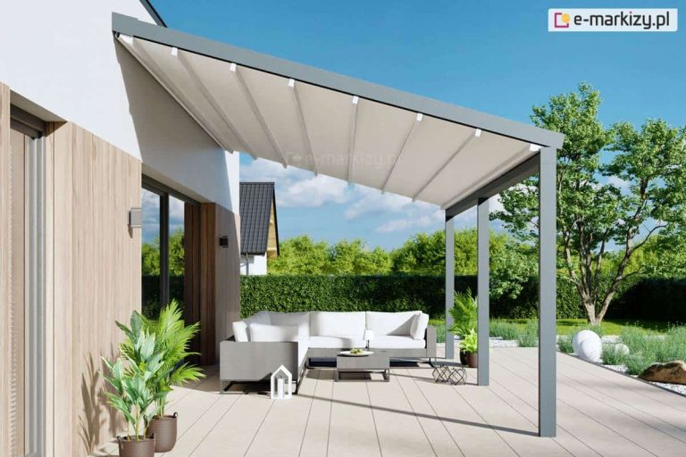 Nowoczesna pergola przyścienna Moderno o konstrukcji z profili aluminiowych w kolorze antracytowym, zamontowana nad tarasem wyłożonym jasnymi płytami. Zadaszenie stanowi segmentowa tkanina w kolorze kremowym, która jest całkowicie rozwinięta i zamontowana pod skosem na prowadnicach, co zapewnia ochronę przed słońcem i opadami. Pod pergolą znajduje się strefa wypoczynkowa z szarym narożnikiem ogrodowym z białymi poduszkami, małym stolikiem kawowym oraz roślinami w donicach. Konstrukcja przylega do elewacji budynku łączącej biały tynk z pionowymi panelami o strukturze jasnego drewna, a w tle widoczny jest zielony żywopłot oraz drzewa w pełnym słońcu.