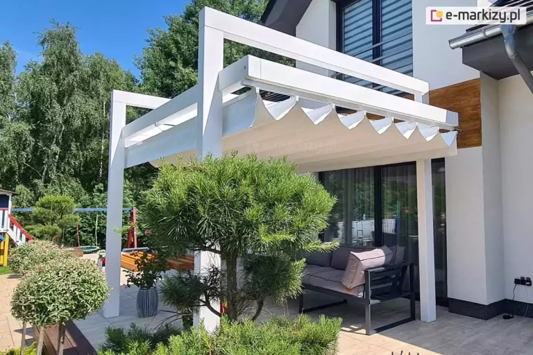 Pergola drewniana kolumnowa Moza w białym kolorze konstrukcji z rozsuniętym systemem zadaszenia tkaninowego. Jasna tkanina dachu rzymskiego jest rozciągnięta pod konstrukcją, tworząc charakterystyczne, głębokie i miękko opadające fałdy w kształcie litery V, które są podwieszone na poziomych prowadnicach. Konstrukcja o białym wykończeniu kontrastuje z ciemnymi profilami okiennymi budynku oraz grafitowym zestawem mebli ogrodowych ustawionych pod zadaszeniem. Na tarasie z jasnych płyt gresowych widoczne jest nowoczesne wyposażenie, w tym sofa z poduszkami oraz rośliny doniczkowe w minimalistycznych, szarych pojemnikach. Na pierwszym planie dominuje dekoracyjnie przycięta sosna oraz krzewy na pniu, rosnące w pasie wysypanym białym grysem. Wzdłuż krawędzi tarasu rośnie lawenda oraz inne niskie krzewy ozdobne. Budynek posiada białą elewację z wstawką z desek elewacyjnych w kolorze ciepłego drewna, co koresponduje z naturalnymi elementami ogrodu. W tle, za konstrukcją pergoli, widoczna jest ściana wysokich, zielonych drzew liściastych oraz fragment bocznej ściany budynku z metalową rynną i osprzętem elektrycznym przy ziemi.