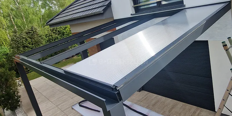 Zdjęcie prezentuje proces montażu pierwszej z czterech płyt poliwęglanowych na aluminiowym szkielecie zadaszenia tarasowego w kolorze antracytowym. Przezroczysta płyta o liniowej strukturze komorowej jest już osadzona w bocznych profilach montażowych i przymocowana do krokwi, natomiast pozostała część konstrukcji ukazuje puste przestrzenie przygotowane pod kolejne arkusze. Całość zadaszenia jest zamontowana do białej elewacji piętrowego budynku, tuż nad ciemną roletą zewnętrzną i oknem z barierką. W dolnej części ujęcia widoczny jest taras wyłożony jasnobrązowymi płytkami ceramicznymi, a w tle gęsta, zielona ściana drzew i krzewów ogrodowych pod jasnym niebem.