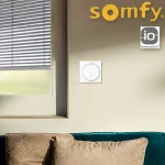 Pilot z ramką Amy 2 Somfy IO zamontowany obok okna wyposażonego w białą żaluzje fasadową