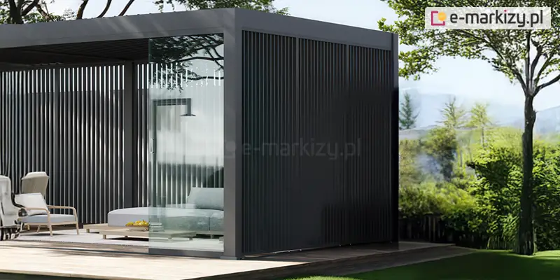 Zdjęcie przedstawia wizualizację nowoczesnej, rozbudowanej pergoli tarasowej w kolorze antracytowym, wkomponowanej w słoneczny ogród. Konstrukcja o płaskim dachu charakteryzuje się pełną zabudową ścian bocznych za pomocą pionowych paneli dekoracyjnych. Panele te składają się z gęsto rozmieszczonych, pionowych listew, które tworzą efekt ażurowej ściany, zapewniając prywatność przy jednoczesnym zachowaniu przewiewności. Frontowa część pergoli jest wyposażona w system przesuwnych paneli szklanych; jeden z nich jest częściowo odsunięty, ukazując wnętrze strefy wypoczynkowej. Wewnątrz pergoli znajdują się jasnoszare meble ogrodowe: niska sofa oraz fotele o minimalistycznym designie. Przed sofą stoi niski stolik kawowy, na którym umieszczono misę z owocami i książki. Całość konstrukcji posadowiona jest na drewnianym podeście tarasowym. Tło wizualizacji stanowi bujna, zielona roślinność z wyraźnie zarysowanymi drzewami liściastymi oraz trawnikiem, skąpana w naturalnym świetle dziennym.