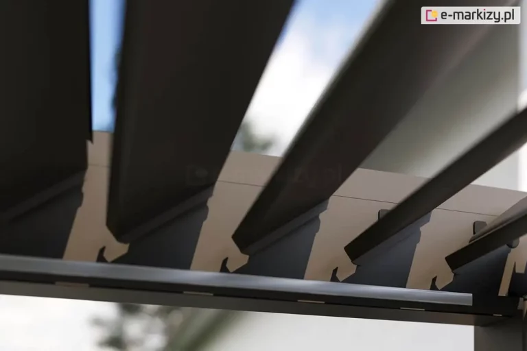 Widok z dołu na detal mechanizmu obrotowego bioklimatycznych piór dachowych w mocno oświetlonej słońcem, antracytowej pergoli Marlo. Zdjęcie przedstawia fragment bocznej szyny prowadzącej, w której zamocowane są końce aluminiowych lamel. Lamele są ustawione pod ostrym kątem w pozycji otwartej, co pozwala na prześwit jasnego nieba i rozproszonego światła pomiędzy poszczególnymi profilami. Każde pióro dachowe posiada charakterystyczne wycięcie montażowe u nasady, umożliwiające jego rotację względem ramy nośnej. Konstrukcja wykonana jest z matowego aluminium w kolorze antracytowym, a na krawędzi dolnej ramy widoczna jest biała listwa zintegrowanego oświetlenia LED. W tle, poza zasięgiem ostrości, widoczne są zarysy zielonych drzew oraz jasna elewacja budynku.