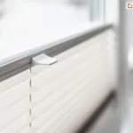 Zdjęcie prezentuje techniczne zbliżenie na górną krawędź systemu przesłony okiennej, jakim jest żaluzja plisowana w jasnym kolorze. Plisa na okno posiada materiał o strukturze drobnej, poziomej harmonijki, która w górnej części jest połączona ze smukłym profilem sterującym w kolorze jasnoszarym. Na środku profilu zamontowany jest ergonomiczny, biały uchwyt o lekko zakrzywionym kształcie, służący do płynnego podnoszenia i opuszczania żaluzji. Mechanizm jest osadzony na pionowych sznurkach prowadzących, które przechodzą przez precyzyjne otwory w tkaninie, zapewniając jej stabilne przyleganie do szyby. Całość zamontowana jest na białej ramie okiennej, widocznej w perspektywie bocznej. Tło za oknem oraz dalsze elementy ramy są mocno rozmyte, co tworzy efekt głębi ostrości i skupia uwagę na detalu uchwytu oraz fakturze materiału. Oświetlenie jest miękkie i naturalne, podkreślając czystość formy i nowoczesny design akcesoriów okiennych.