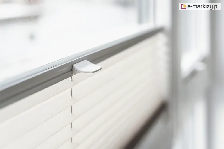 Zdjęcie prezentuje techniczne zbliżenie na górną krawędź systemu przesłony okiennej, jakim jest żaluzja plisowana w jasnym kolorze. Plisa na okno posiada materiał o strukturze drobnej, poziomej harmonijki, która w górnej części jest połączona ze smukłym profilem sterującym w kolorze jasnoszarym. Na środku profilu zamontowany jest ergonomiczny, biały uchwyt o lekko zakrzywionym kształcie, służący do płynnego podnoszenia i opuszczania żaluzji. Mechanizm jest osadzony na pionowych sznurkach prowadzących, które przechodzą przez precyzyjne otwory w tkaninie, zapewniając jej stabilne przyleganie do szyby. Całość zamontowana jest na białej ramie okiennej, widocznej w perspektywie bocznej. Tło za oknem oraz dalsze elementy ramy są mocno rozmyte, co tworzy efekt głębi ostrości i skupia uwagę na detalu uchwytu oraz fakturze materiału. Oświetlenie jest miękkie i naturalne, podkreślając czystość formy i nowoczesny design akcesoriów okiennych.