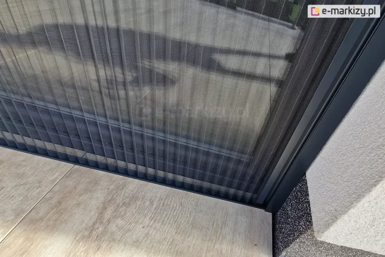 W pełni rozwinięta szara siatka moskitiery plisowanej Top Zag układająca się w charakterystyczne fale zamocowana do ściany wnęki okiennej oraz do płytek tarasowych o drewnianym wzorze.