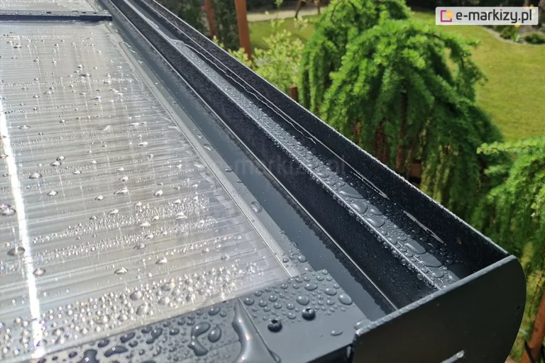 Zbliżenie rynny ukrytej w belce przedniej zadaszenia z poliwęglanu oraz jej połączenia z płytami poliwęglanowymi. Całość pokryta wodą, która zebrała się w większe i mniejsze krople na dachu i konstrukcji. Obok zadaszenia widoczne zielone krzewy i świeżo skoszony trawnik.