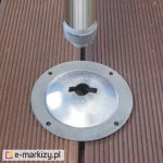 Okrągła metalowa stopka montażowa z centralnym klinowym wycięciem i czterema otworami na śruby osadzona w deskowanej nawierzchni tarasu, nad nią zakończenie aluminiowego słupka.