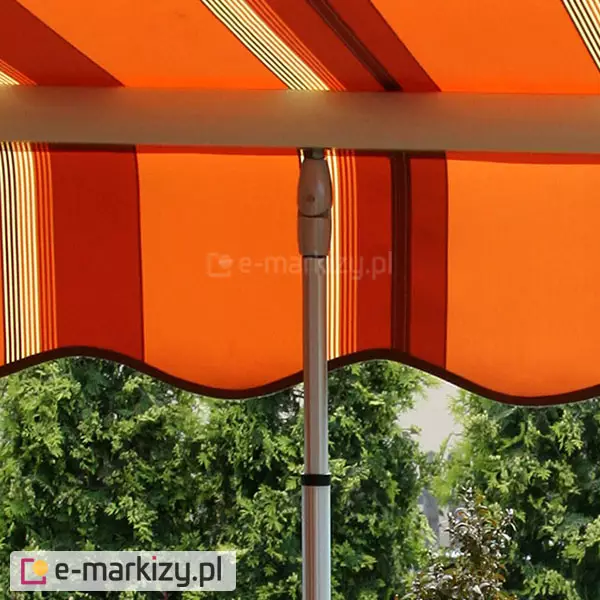 Belka przednia markizy tarasowej MOL z tkaniną w pomarańczowe, czerwone, brązowe i białe pasy z umieszczoną w niej podporą teleskopową. Pod falowaną falbaną z ciemnobrązową lamówka widoczne zielone krzewy.