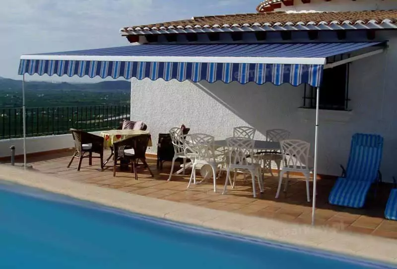 Taras przy basenie wyłożony brązowo-pomarańczowymi kaflami, na który ustawiono dwa stoły: wiklinowy nakryty ceratą w żółtym kolorze i czterema krzesłami oraz biały z sześcioma krzesłami i dwa leżaki z poduszkami w niebieskie pasy. Całość zacieniona została markizą o białej konstrukcji z poszyciem w pasy w kilku odcieniach niebieskiego, z belką przednią wspartą na dwóch podporach teleskopowych. Markizę zamocowano do białej ściany domku pod daszkiem wyłożonym dachówką. W tle za barierką tarasową widoczna panorama okolicy pełna zieleni.