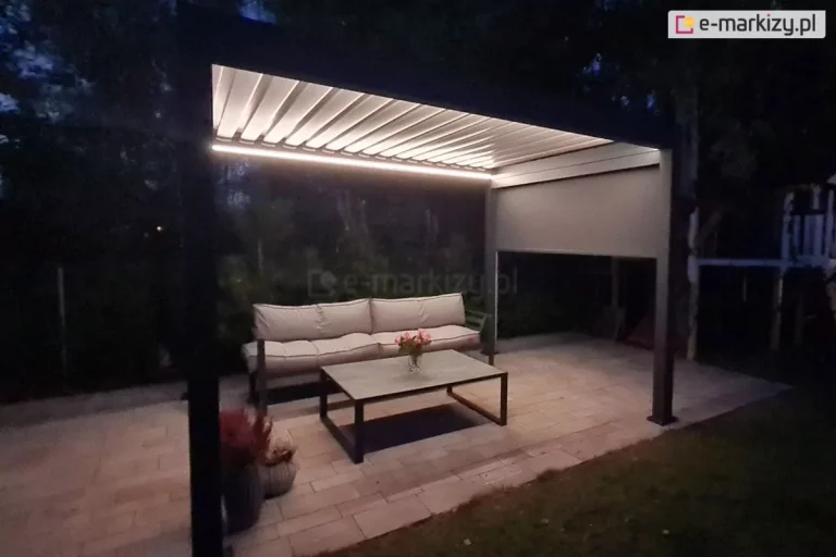 Pergola Essential Lamelowa Wolnostojąca Galeria Zdjęć: Nowoczesna pergola tarasowa o konstrukcji aluminiowej w kolorze ciemnego antracytu, sfotografowana po zmroku z włączonym zintegrowanym oświetleniem LED. Zadaszenie pergoli składa się z ruchomych lameli w kolorze białym, które są ustawione w pozycji poziomej, a pod nimi, wzdłuż wewnętrznych krawędzi ramy, zamontowano liniowe taśmy LED emitujące ciepłe, jasne światło. Na jednej z bocznych ścian konstrukcji widoczna jest częściowo rozwinięta roleta zewnętrzna typu screen w kolorze szarym, która zasłania górną połowę prześwitu między słupami. Pod pergolą, na podłożu wyłożonym jasną kostką brukową, zaaranżowano strefę wypoczynkową z wygodną sofą ogrodową z beżowymi poduszkami oraz niskim stolikiem kawowym z ciemnym stelażem, na którym stoi wazon z kwiatami. W otoczeniu konstrukcji panuje półmrok, a w tle zarysowują się ciemne sylwetki drzew i gęstej roślinności ogrodowej. Całość prezentuje funkcjonalne wykorzystanie oświetlenia tarasowego w architekturze ogrodowej, tworząc przytulną atmosferę do odpoczynku wieczorową porą.
