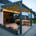 Pergola przyścienna Elegancy Tarasola z elektrycznie rozsuwanym dachem tkaninowym i wbudowanym oświetleniem LED tworzy elegancką, wieczorną strefę wypoczynkową na tarasie. Pod zadaszeniem ustawiono drewniany zestaw mebli oraz grill, a całość otacza zadbany ogród i donice z kolorowymi kwiatami.