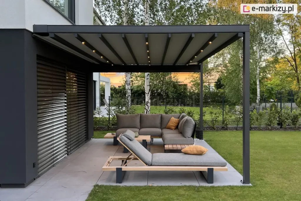 Taras ogrodowy z nowoczesnym zadaszeniem typu pergola. Konstrukcja składa się z grafitowych, metalowych słupów i belkowanego dachu z pokryciem z tkaniny o jasnoszarym kolorze. Pomiędzy pasami tkaniny widoczne belki przeciwwietrzne z wbudowanymi punktami świetlnymi, aktualnie podświetlającymi przestrzeń pod pergolą. W centrum kadru znajduje się komplet mebli wypoczynkowych: narożna sofa i leżanka o minimalistycznym designie, wykonane z jasnoszarej tkaniny na drewniano-metalowej ramie. Na siedziskach ułożono poduszki dekoracyjne w kolorze musztardowym. Zestaw uzupełnia niski stolik kawowy. Meble ustawione są na tarasie wyłożonym wielkoformatowymi, szarymi płytkami. Za tarasem widoczny jest trawnik i niski żywopłot. W dalszym planie znajdują się drzewa liściaste – brzozy i inne gatunki – oraz nowoczesna zabudowa mieszkalna. Po lewej stronie zdjęcia widać elewację domu z ciemnymi poziomymi żaluzjami osłaniającymi duże przeszklenie.