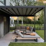 Taras ogrodowy z nowoczesnym zadaszeniem typu pergola. Konstrukcja składa się z grafitowych, metalowych słupów i belkowanego dachu z pokryciem z tkaniny o jasnoszarym kolorze. Pomiędzy pasami tkaniny widoczne belki przeciwwietrzne z wbudowanymi punktami świetlnymi, aktualnie podświetlającymi przestrzeń pod pergolą. W centrum kadru znajduje się komplet mebli wypoczynkowych: narożna sofa i leżanka o minimalistycznym designie, wykonane z jasnoszarej tkaniny na drewniano-metalowej ramie. Na siedziskach ułożono poduszki dekoracyjne w kolorze musztardowym. Zestaw uzupełnia niski stolik kawowy. Meble ustawione są na tarasie wyłożonym wielkoformatowymi, szarymi płytkami. Za tarasem widoczny jest trawnik i niski żywopłot. W dalszym planie znajdują się drzewa liściaste – brzozy i inne gatunki – oraz nowoczesna zabudowa mieszkalna. Po lewej stronie zdjęcia widać elewację domu z ciemnymi poziomymi żaluzjami osłaniającymi duże przeszklenie.