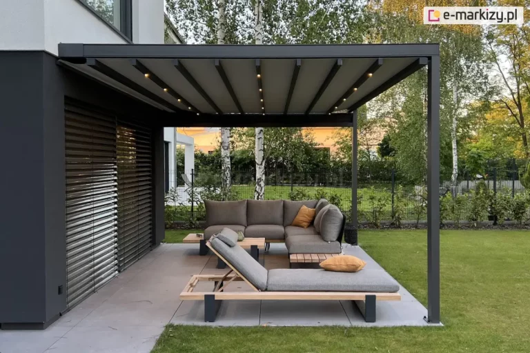 Taras ogrodowy z nowoczesnym zadaszeniem typu pergola. Konstrukcja składa się z grafitowych, metalowych słupów i belkowanego dachu z pokryciem z tkaniny o jasnoszarym kolorze. Pomiędzy pasami tkaniny widoczne belki przeciwwietrzne z wbudowanymi punktami świetlnymi, aktualnie podświetlającymi przestrzeń pod pergolą. W centrum kadru znajduje się komplet mebli wypoczynkowych: narożna sofa i leżanka o minimalistycznym designie, wykonane z jasnoszarej tkaniny na drewniano-metalowej ramie. Na siedziskach ułożono poduszki dekoracyjne w kolorze musztardowym. Zestaw uzupełnia niski stolik kawowy. Meble ustawione są na tarasie wyłożonym wielkoformatowymi, szarymi płytkami. Za tarasem widoczny jest trawnik i niski żywopłot. W dalszym planie znajdują się drzewa liściaste – brzozy i inne gatunki – oraz nowoczesna zabudowa mieszkalna. Po lewej stronie zdjęcia widać elewację domu z ciemnymi poziomymi żaluzjami osłaniającymi duże przeszklenie.
