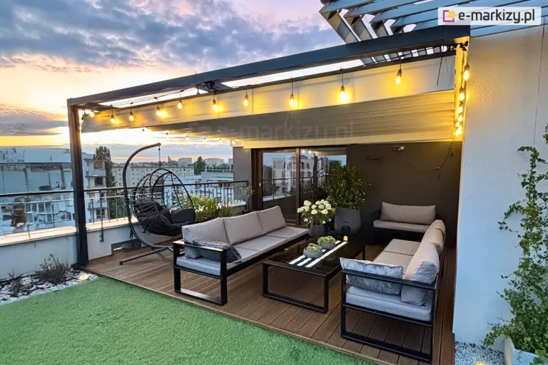 Nowoczesna strefa relaksu na tarasie apartamentu, zorganizowana pod czarną, metalową konstrukcją pergoli z zamontowanym jasnym dachem rzymskim Supro. Tkanina zadaszenia jest rozciągnięta poziomo, a wzdłuż krawędzi konstrukcji zawieszono girlandę z żarówkami dekoracyjnymi, które emitują intensywne, ciepłe światło. Na tarasie znajduje się zestaw mebli wypoczynkowych składający się z dwóch szarych sof z miękkimi poduchami oraz czarnego, niskiego stolika kawowego ze szklanym blatem. Po lewej stronie widoczny jest wiszący fotel typu kokon na stabilnym stelażu. Podłoga tarasu jest podzielona na dwie strefy: wypoczynkową wyłożoną brązowymi deskami kompozytowymi oraz rekreacyjną z nawierzchnią ze sztucznej trawy. Przy krawędziach tarasu umieszczono donice z krzewami oraz białymi kwiatami, a za niską balustradą rozciąga się widok na panoramę miasta z budynkami mieszkalnymi pod wieczornym, lekko zachmurzonym niebem w odcieniach błękitu i różu.