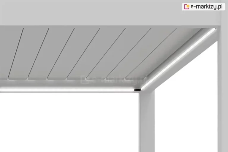 Zbliżenie na wewnętrzny narożnik białej pergoli Marlo, prezentujące zintegrowane oświetlenie LED wmontowane w ramę konstrukcji. Zdjęcie ukazuje punkt styku poziomej belki dachowej z pionowym słupem, gdzie wzdłuż wewnętrznych krawędzi profilu biegną dwie prostopadłe do siebie taśmy LED, emitujące jasne, białe światło o ciągłej linii. Powyżej linii światła widoczne są aluminiowe lamele dachowe w kolorze białym, ułożone równolegle obok siebie w pozycji zamkniętej. Całość konstrukcji jest utrzymana w jednolitej, czystej bieli o matowym wykończeniu, a minimalistyczny sposób montażu oświetlenia sprawia, że źródło światła jest zlicowane z powierzchnią profilu. Detal ten podkreśla nowoczesny design i funkcjonalność systemu zadaszenia, zapewniając estetyczne doświetlenie przestrzeni pod pergolą.