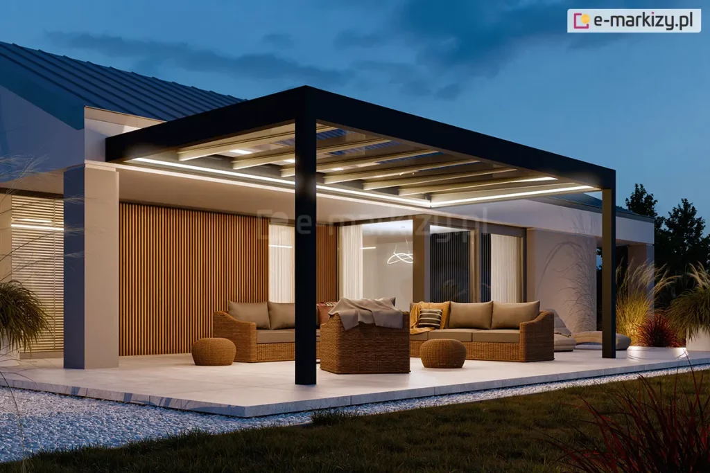 Wieczorna ekspozycja nowoczesnego zadaszenia tarasowego Spekta Glass, zintegrowanego z bryłą budynku jednorodzinnego o jasnej elewacji. Konstrukcja nośna została wykonana z grubych profili aluminiowych w kolorze antracytowym, które tworzą geometryczną ramę wspartą na pionowych słupach. System został wyposażony w zintegrowane oświetlenie LED w formie intensywnych, liniowych pasów świetlnych zamontowanych wzdłuż ramy obwodowej, które oświetlają całą strefę wypoczynku. Wewnątrz przeszklonej strefy zaaranżowano komfortowe miejsce do relaksu z kompletem mebli z technorattanu: dwiema sofami, fotelem z brązowymi poduchami oraz niskimi, okrągłymi pufami. Ściana budynku pod zadaszeniem została wykończona dekoracyjnymi, pionowymi lamelami drewnianymi, co podkreśla nowoczesny design. Podłoże stanowią wielkoformatowe płyty tarasowe w jasnym odcieniu, otoczone pasem białego gresu i ciemnym trawnikiem. Całość kompozycji, dopełniona przez nastrojowe oświetlenie ogrodowe i roślinność w tle, tworzy luksusową i spójną przestrzeń wypoczynku.