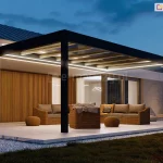 Wieczorna ekspozycja nowoczesnego zadaszenia tarasowego Spekta Glass, zintegrowanego z bryłą budynku jednorodzinnego o jasnej elewacji. Konstrukcja nośna została wykonana z grubych profili aluminiowych w kolorze antracytowym, które tworzą geometryczną ramę wspartą na pionowych słupach. System został wyposażony w zintegrowane oświetlenie LED w formie intensywnych, liniowych pasów świetlnych zamontowanych wzdłuż ramy obwodowej, które oświetlają całą strefę wypoczynku. Wewnątrz przeszklonej strefy zaaranżowano komfortowe miejsce do relaksu z kompletem mebli z technorattanu: dwiema sofami, fotelem z brązowymi poduchami oraz niskimi, okrągłymi pufami. Ściana budynku pod zadaszeniem została wykończona dekoracyjnymi, pionowymi lamelami drewnianymi, co podkreśla nowoczesny design. Podłoże stanowią wielkoformatowe płyty tarasowe w jasnym odcieniu, otoczone pasem białego gresu i ciemnym trawnikiem. Całość kompozycji, dopełniona przez nastrojowe oświetlenie ogrodowe i roślinność w tle, tworzy luksusową i spójną przestrzeń wypoczynku.