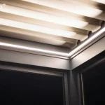 Zbliżenie na detal techniczny i oświetlenie nowoczesnej pergoli Essential tarasola po zmroku. Zdjęcie przedstawia górny narożnik konstrukcji, gdzie łączą się ciemnoantracytowe profile aluminiowe ramy. Wewnątrz profili konstrukcyjnych zintegrowano liniowe oświetlenie LED w formie białych listew świecących jasnym, chłodnym światłem, które biegną wzdłuż krawędzi zadaszenia. Nad oświetleniem widoczne są białe lamele dachowe, które od spodu są mocno doświetlone przez system LED, co tworzy efektowne pasy światła i cienia. Poniżej linii oświetlenia widoczna jest ciemna, jednolita płaszczyzna opuszczonej rolety lub wypełnienia bocznego, kontrastująca z jasnymi elementami świetlnymi konstrukcji.