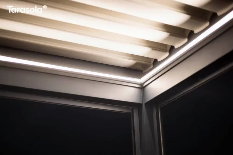 Zbliżenie na detal techniczny i oświetlenie nowoczesnej pergoli Essential tarasola po zmroku. Zdjęcie przedstawia górny narożnik konstrukcji, gdzie łączą się ciemnoantracytowe profile aluminiowe ramy. Wewnątrz profili konstrukcyjnych zintegrowano liniowe oświetlenie LED w formie białych listew świecących jasnym, chłodnym światłem, które biegną wzdłuż krawędzi zadaszenia. Nad oświetleniem widoczne są białe lamele dachowe, które od spodu są mocno doświetlone przez system LED, co tworzy efektowne pasy światła i cienia. Poniżej linii oświetlenia widoczna jest ciemna, jednolita płaszczyzna opuszczonej rolety lub wypełnienia bocznego, kontrastująca z jasnymi elementami świetlnymi konstrukcji.