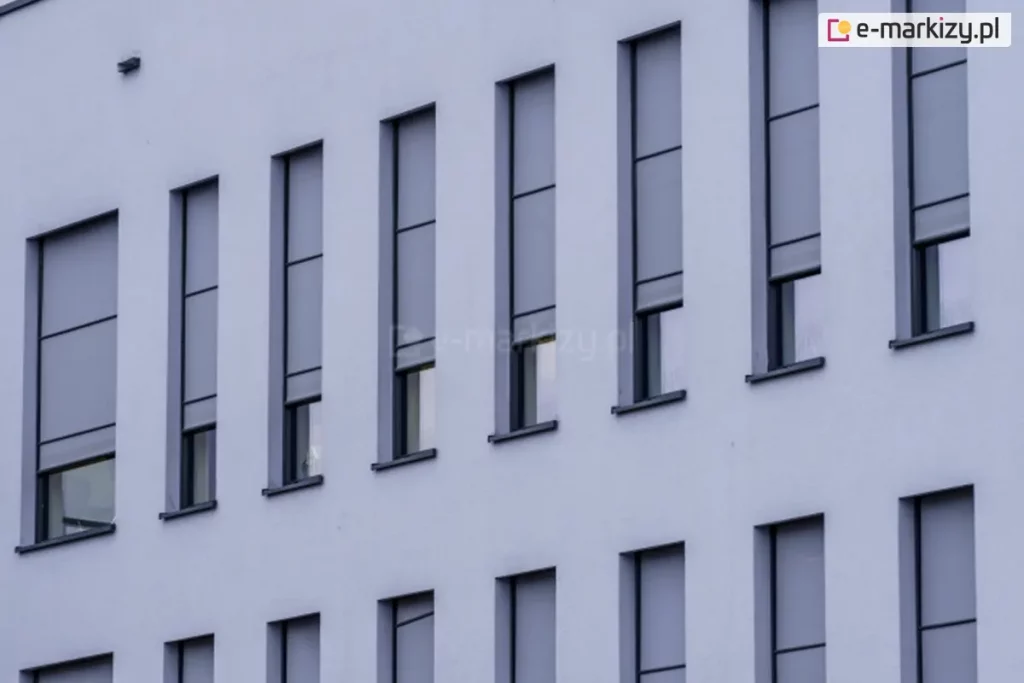 Elewacja nowoczesnego budynku z wąskimi, pionowymi oknami, na których zamontowano zewnętrzne rolety screenowe. Rolety wykonane są z półprzezroczystej tkaniny w kolorze szarym i częściowo zasłaniają przeszklenia, pozostawiając dolne części okien odkryte. System prowadnic bocznych zapewnia stabilne prowadzenie tkaniny, co sugeruje zastosowanie rozwiązania typu ZIP. Kasety rolet są wbudowane i licują z elewacją, co zapewnia estetyczny i minimalistyczny wygląd fasady.