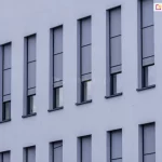 Elewacja nowoczesnego budynku z wąskimi, pionowymi oknami, na których zamontowano zewnętrzne rolety screenowe. Rolety wykonane są z półprzezroczystej tkaniny w kolorze szarym i częściowo zasłaniają przeszklenia, pozostawiając dolne części okien odkryte. System prowadnic bocznych zapewnia stabilne prowadzenie tkaniny, co sugeruje zastosowanie rozwiązania typu ZIP. Kasety rolet są wbudowane i licują z elewacją, co zapewnia estetyczny i minimalistyczny wygląd fasady.