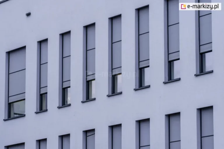 Elewacja nowoczesnego budynku z wąskimi, pionowymi oknami, na których zamontowano zewnętrzne rolety screenowe. Rolety wykonane są z półprzezroczystej tkaniny w kolorze szarym i częściowo zasłaniają przeszklenia, pozostawiając dolne części okien odkryte. System prowadnic bocznych zapewnia stabilne prowadzenie tkaniny, co sugeruje zastosowanie rozwiązania typu ZIP. Kasety rolet są wbudowane i licują z elewacją, co zapewnia estetyczny i minimalistyczny wygląd fasady.