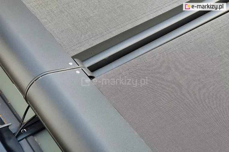 Zbliżenie na kasety dwóch modułów systemu Veranda w kolorze antracytowym. Zdjęcie prezentuje miejsce styku dwóch modułów zaokrąglonej, aluminiowej obudowy malowanej proszkowo. Z kasety wyprowadzone są prowadnice, pomiędzy którymi znajduje się niewielka szczelina. Przy każdej z prowadnic widoczna jest ciemnoszara tkanina akrylowa. W lewym dolnym rogu widoczne są czarne przewody elektryczne doprowadzające zasilanie do silnika markizy oraz fragment gumowej uszczelki. Tkanina jest mocno napięta, co świadczy o sprawnym działaniu systemu naciągu. Konstrukcja jest zamontowana na szklanym zadaszeniu, którego krawędź jest widoczna pod kasetą.