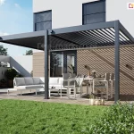 Wizualizacja nowoczesnej, dwumodułowej pergoli tarasowej z dachem lamelowym model Rubio Soft, zamontowanej przy elewacji nowoczesnego piętrowego domu o białej fasadzie. Konstrukcja pergoli jest wykonana z aluminium w kolorze antracytowym i opiera się na trzech pionowych słupach o kwadratowym przekroju, które są zakotwione w drewnianym tarasie. Zadaszenie składa się z dwóch sekcji ruchomych lameli – w lewym module lamele są mocno przymknięte, natomiast w prawym module są one otwarte, wpuszczając światło słoneczne i tworząc charakterystyczny wzór pasów światła i cienia na elewacji budynku. Przy lewym i środkowym słupie zamontowane są manualne korby do regulacji kąta nachylenia lameli. Pod pergolą znajduje się strefa wypoczynkowa z dużym, jasnym narożnikiem ogrodowym oraz strefa jadalniana z nowoczesnym stołem i sześcioma krzesłami w kolorze białym. Na stole widoczna jest zastawa, a obok stoją dekoracyjne lampiony. W tle widać fragment zielonego trawnika, niskie nasadzenia w białych donicach oraz nowoczesne ogrodzenie. Elewacja parteru pod pergolą wykończona jest poziomymi deskami w naturalnym odcieniu drewna, z zamontowanymi czarnymi kinkietami zewnętrznymi. Duże przeszklenia i drzwi tarasowe w ciemnych ramach prowadzą do wnętrza budynku.