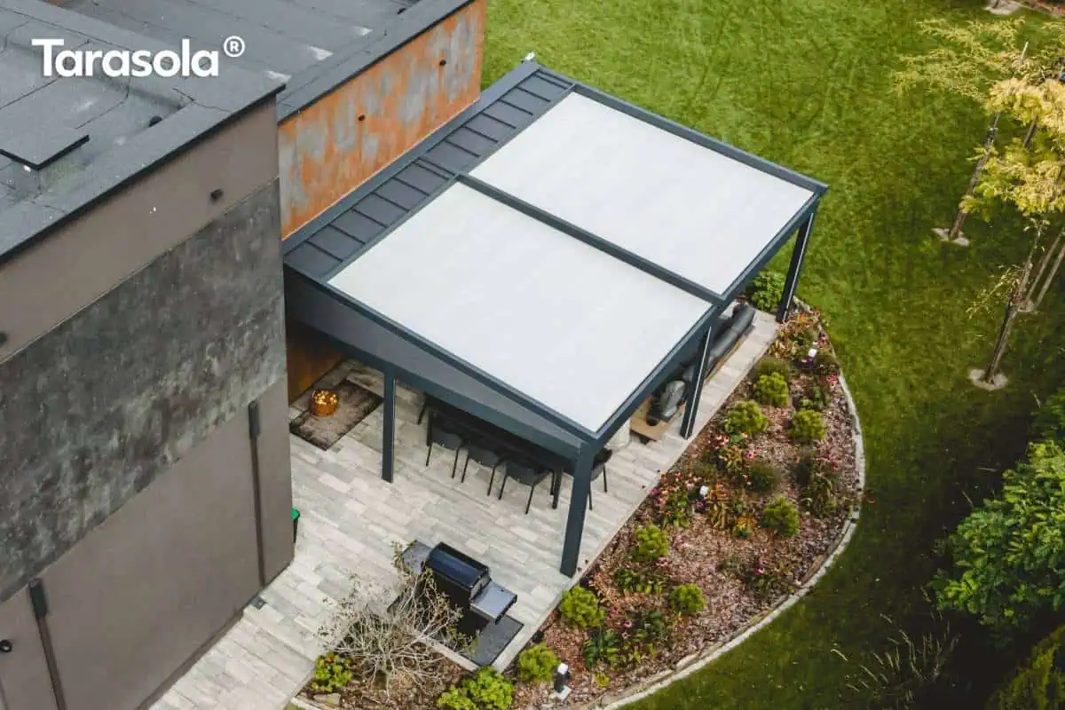 Widok z lotu ptaka na nowoczesną, podwójną pergolę tarasową o konstrukcji aluminiowej w ciemnym kolorze antracytowym, zamontowaną przy elewacji budynku o surowym, industrialnym wykończeniu. Zadaszenie składa się z dwóch modułów z jasną, napinaną tkaniną chroniącą przed słońcem i deszczem, z widocznym systemem odprowadzania wody w postaci systemowych daszków osłonowych przy ścianie. Pod pergolą, na tarasie wyłożonym jasnymi płytami gresowymi, zaaranżowano strefę jadalnianą z długim stołem i krzesłami oraz strefę wypoczynkową z ciemną sofą ogrodową. Obok tarasu znajduje się profesjonalny grill gazowy, a całość otacza starannie zaprojektowana roślinność ogrodowa z niskimi krzewami i korą, oddzielona od zielonego trawnika półkolistym obrzeżem. Konstrukcja pergoli przylega do ściany pokrytej panelami o efekcie rdzy (corten) oraz szarym tynkiem strukturalnym, co podkreśla nowoczesny charakter inwestycji.