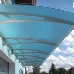 Daszek nad drzwi Elegant łączony z wielu modułów zamontowany na elewacji budynku przemysłowego lub użyteczności publicznej, tworzący długi ciąg ochronny nad wejściami. Konstrukcja składa się z wielu połączonych segmentów o łukowym kształcie, wspartych na stalowych wspornikach o szarym kolorze z charakterystycznymi geometrycznymi wycięciami. Poszycie wykonano z akrylu o niebieskim zabarwieniu, który skutecznie rozprasza światło słoneczne, nadając zadaszeniu nowoczesny wygląd. Daszki osłaniają białą elewację o tynku strukturalnym, na której widoczna jest tabliczka informacyjna „Tor 2” nad jednym z wejść. W tle rozpościera się dynamiczne, letnie niebo z białymi, pierzastymi chmurami (cumulusami), a w dolnej części kadru widać wierzchołki drzew iglastych i liściastych oraz fragmenty zabudowy miejskiej. Perspektywa zdjęcia z dołu podkreśla dużą powierzchnię całkowitą zadaszenia oraz precyzję łączenia poszczególnych modułów.