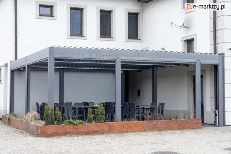 Duża, modułowa pergola tarasowa Sb400 w kolorze antracytowym, zainstalowana przy jasnej elewacji budynku jako zadaszenie ogródka restauracyjnego. Konstrukcja składa się z trzech segmentów, z których dwa są wyposażone w przesłony boczne (screeny) w kolorze szarym, zapewniające ochronę przed wiatrem i słońcem. Pod pergolą ustawiono liczne zestawy mebli gastronomicznych – czarne stoły i dopasowane do nich krzesła z tworzywa. Całość odgrodzona jest od placu z kostki brukowej długą, niską donicą wykonaną z drewna o ciepłym odcieniu, w której posadzono małe iglaki i ozdobne trawy. Na ścianie budynku powyżej pergoli widoczne są klasyczne okna w brązowych ramach oraz antena satelitarna. Zdjęcie prezentuje profesjonalną zabudowę przestrzeni komercyjnej, łączącą funkcję ochrony przed pogodą z estetycznym wydzieleniem strefy dla gości.