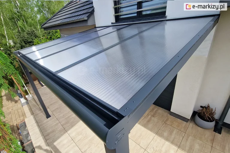 Zadaszenie tarasu wykonane z aluminiowej konstrukcji w kolorze antracytowym oraz płyt poliwęglanu komorowego 16mm o przejrzystej barwie. Nad zadaszeniem widoczne okno z balkonem francuskim oraz spadzisty dach pokryty dachówką. Pod zadaszeniem taras wyłożony płytkami w odcieniach jasnego brązu imitujące strukturę drewna. Wokół liczne zielone drzewa.