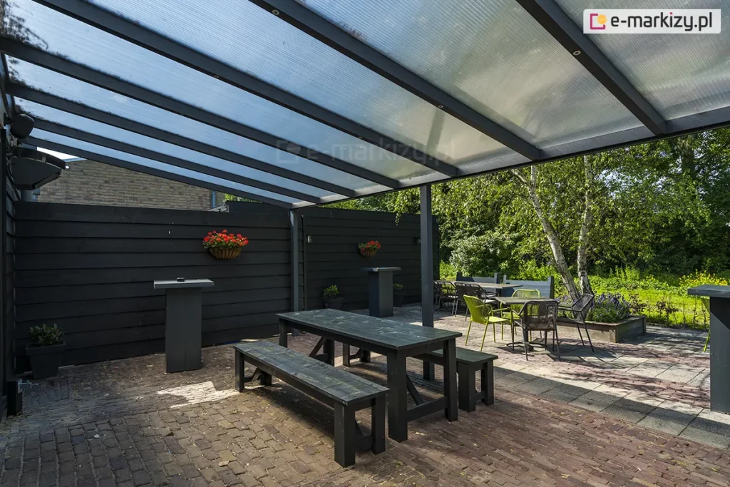 Zdjęcie prezentuje przestronną strefę rekreacyjną pod zadaszeniem tarasowym Nebbiolo marki Deponti, pokrytym płytami z przezroczystego poliwęglanu komorowego. Konstrukcja dachu opiera się na gęsto rozstawionych krokwiach z aluminium w kolorze antracytowym, które są podtrzymywane przez poprzeczną belkę i pionowy słup nośny. Pod zadaszeniem znajduje się zestaw mebli ogrodowych, w tym długi, czarny stół z dwiema ławami wykonanymi z drewna malowanego na ciemny kolor oraz mniejsze stoliki kawowe z krzesłami w kolorze limonkowym i czarnym. Taras jest ograniczony wysokim, pełnym płotem z poziomych desek w kolorze czarnym, na którym zawieszono donice z czerwonymi kwiatami. Nawierzchnia tarasu została wykonana z postarzanej kostki brukowej w odcieniach brązu i szarości. W tle widoczna jest gęsta roślinność ogrodowa z dominującą brzozą o jasnej korze oraz fragmenty trawnika pod słonecznym niebem.