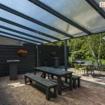 Zdjęcie prezentuje przestronną strefę rekreacyjną pod zadaszeniem tarasowym Nebbiolo marki Deponti, pokrytym płytami z przezroczystego poliwęglanu komorowego. Konstrukcja dachu opiera się na gęsto rozstawionych krokwiach z aluminium w kolorze antracytowym, które są podtrzymywane przez poprzeczną belkę i pionowy słup nośny. Pod zadaszeniem znajduje się zestaw mebli ogrodowych, w tym długi, czarny stół z dwiema ławami wykonanymi z drewna malowanego na ciemny kolor oraz mniejsze stoliki kawowe z krzesłami w kolorze limonkowym i czarnym. Taras jest ograniczony wysokim, pełnym płotem z poziomych desek w kolorze czarnym, na którym zawieszono donice z czerwonymi kwiatami. Nawierzchnia tarasu została wykonana z postarzanej kostki brukowej w odcieniach brązu i szarości. W tle widoczna jest gęsta roślinność ogrodowa z dominującą brzozą o jasnej korze oraz fragmenty trawnika pod słonecznym niebem.