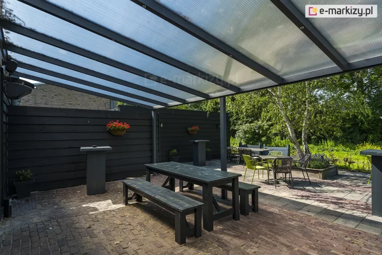 Zdjęcie prezentuje przestronną strefę rekreacyjną pod zadaszeniem tarasowym Nebbiolo marki Deponti, pokrytym płytami z przezroczystego poliwęglanu komorowego. Konstrukcja dachu opiera się na gęsto rozstawionych krokwiach z aluminium w kolorze antracytowym, które są podtrzymywane przez poprzeczną belkę i pionowy słup nośny. Pod zadaszeniem znajduje się zestaw mebli ogrodowych, w tym długi, czarny stół z dwiema ławami wykonanymi z drewna malowanego na ciemny kolor oraz mniejsze stoliki kawowe z krzesłami w kolorze limonkowym i czarnym. Taras jest ograniczony wysokim, pełnym płotem z poziomych desek w kolorze czarnym, na którym zawieszono donice z czerwonymi kwiatami. Nawierzchnia tarasu została wykonana z postarzanej kostki brukowej w odcieniach brązu i szarości. W tle widoczna jest gęsta roślinność ogrodowa z dominującą brzozą o jasnej korze oraz fragmenty trawnika pod słonecznym niebem.