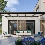 Nowoczesne zadaszenie tarasowe Lunaro w kolorze antracytowym, zamontowane do jasnej elewacji minimalistycznego, piętrowego budynku. Konstrukcja dachu opiera się na poprzecznych aluminiowych krokwiach z wypełnieniem z mlecznych paneli poliwęglanowych, a całość wspierają dwa smukłe, kwadratowe słupy nośne ustawione na brzegach tarasu. Na pierwszym planie widoczne są intensywnie fioletowe kwiaty lawendy, a w tle wysokie drzewa liściaste pod jasnym, pogodnym niebem. Pod zadaszeniem, na jasnej i gładkiej nawierzchni, zaaranżowano strefę relaksu z designerską sofą ozdobioną miętowo-beżowymi poduszkami, dwoma fotelami w podobnym stylu oraz niskim, prostokątnym stolikiem kawowym. Przeszklone wejście do domu zdobią pionowe, drewniane lamele, które pełnią funkcję dekoracyjną i osłonową.