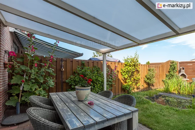 Perspektywiczny widok spod zadaszenia tarasowego na przydomowy ogród, eksponujący detale konstrukcji dachu oraz aranżację strefy jadalnianej. Konstrukcja zadaszenia w kolorze kremowym lub białym posiada dach wykonany z paneli poliwęglanu komorowego, które są przedzielone równoległymi profilami aluminiowymi. Bezpośrednio pod dachem ustawiono duży, prostokątny stół z grubym drewnianym blatem o strukturze postarzanego drewna, na którym stoi ceramiczna donica z czerwonymi kwiatami oraz mała świeca zapachowa w fioletowym kolorze. Przy stole znajdują się cztery głębokie fotele wykonane z technorattanu w odcieniach szarości z zaokrąglonymi oparciami. Po lewej stronie widoczna jest elewacja budynku z czerwonej cegły, przy której rośnie wysoka malwa z licznymi różowymi kwiatami, wspierająca się o brązowy, pionowy płot z drewnianych desek. W głębi ogrodu, za pasem zielonej trawy, znajduje się oczko wodne zabezpieczone siatką, otoczone bujną roślinnością, a po prawej stronie, przy płocie, stoi murowany, szary kominek ogrodowy z daszkiem. Całość kompozycji dopełnia widok bezchmurnego nieba oraz różnorodnych krzewów i pnączy rosnących wzdłuż ogrodzenia.