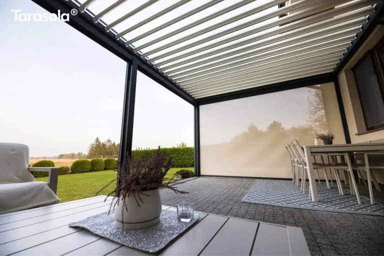 Perspektywa z poziomu stolika kawowego ukazująca wnętrze i konstrukcję dachu Essential tarasola. Na pierwszym planie widoczny jest fragment białego blatu stolika z doniczką z fioletowym wrzosem i szklanym lampionem na srebrnej podkładce. Nad tarasem znajduje się dach z białych lameli aluminiowych, które są otwarte, ukazując fragmenty jasnego nieba. Konstrukcja wsparta jest na solidnych, ciemnych profilach aluminiowych. Jedna ze ścian bocznych jest całkowicie zasłonięta przez dużą, pionową roletę screen w odcieniu beżowym, na której malują się delikatne cienie rzucane przez pobliskie drzewa. Pod ścianą budynku stoi biały stół jadalniany z krzesłami ustawiony na geometrycznym dywanie. Podłoga tarasu wyłożona jest szarą kostką brukową. Przez otwarty bok pergoli widać fragment ogrodu z zielonym trawnikiem, rzędem formowanych krzewów oraz szeroki horyzont.